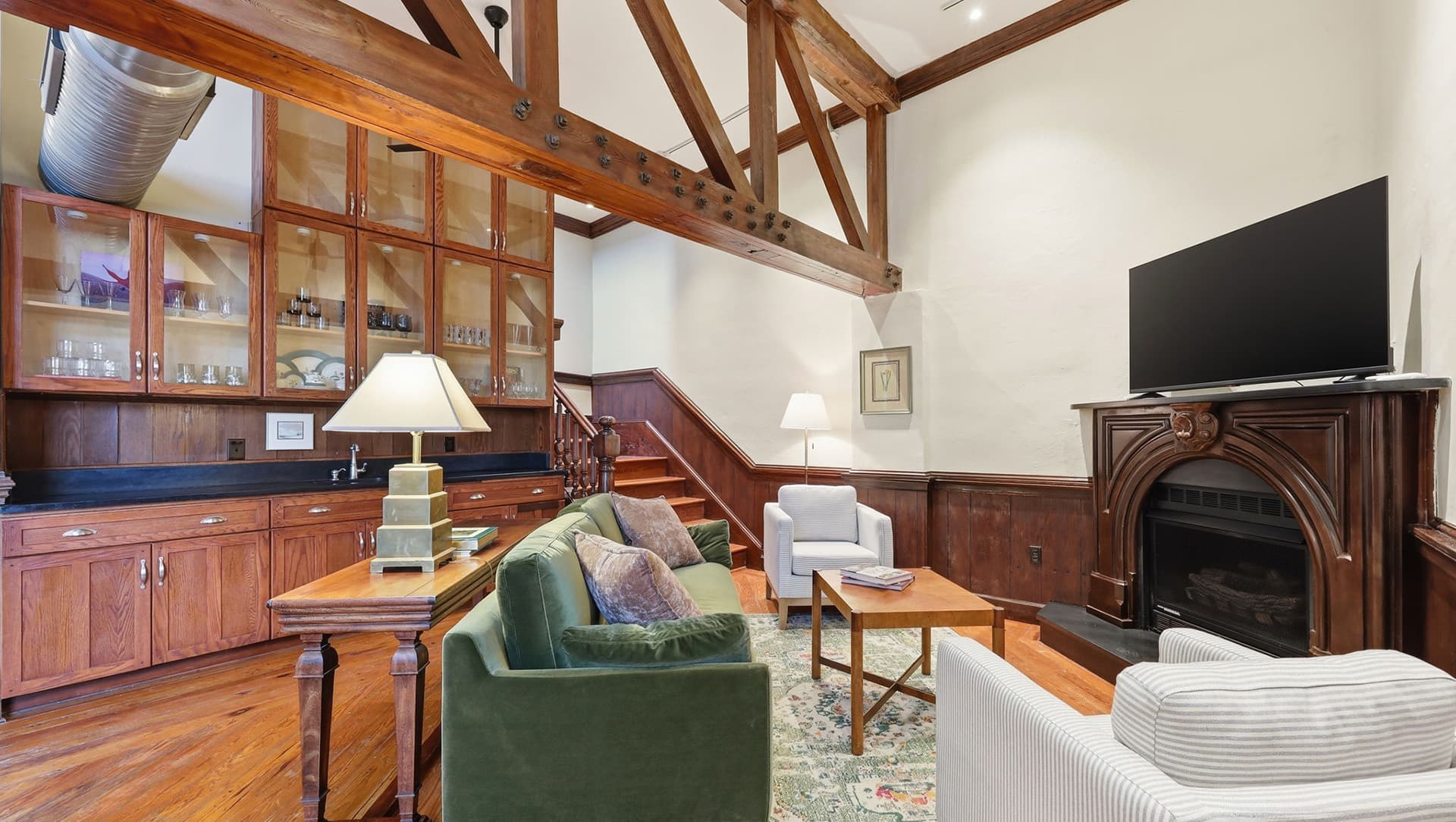 A cozy living room featuring green and white sofas, wooden beams, a fireplace, and a large TV.